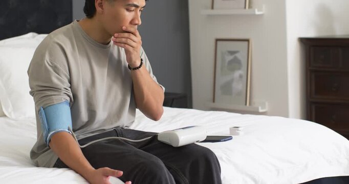 Asian man sitting on bed with blood pressure cuff on left arm, waiting for monitor reading