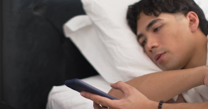Asian man lying on bed, reaching for smartphone while scrolling beside alarm clock 5:38