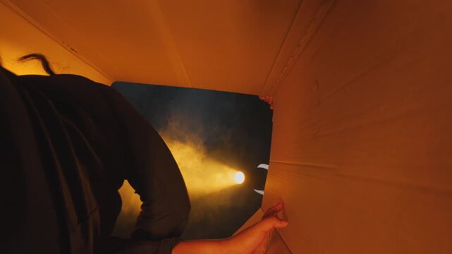 Unique point-of-view from inside a cardboard box as someone closes the top flaps, trapping the viewer inside. Smoke and warm light leak in. Represents kidnapping, moving, or fear.