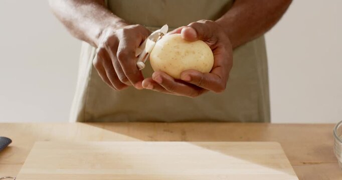 Vertical video: Man wearing apron placing board, peeling potato with Y peeler at counter to cook