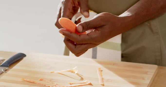 Adult African American man in striped apron prepping meal peeling carrot with peeler on board