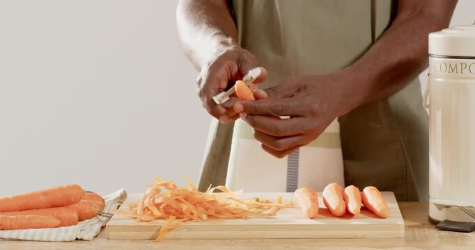 African American man wearing striped apron picking up carrot, peeling with peeler for meal prep