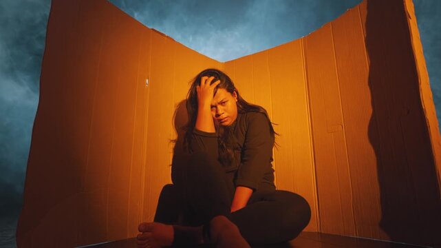 A lonely woman leans her head back against a cardboard wall in a surreal studio setting. Heavy blue smoke surrounds the warm, confined space, visualizing depression, isolation, and feeling trapped.