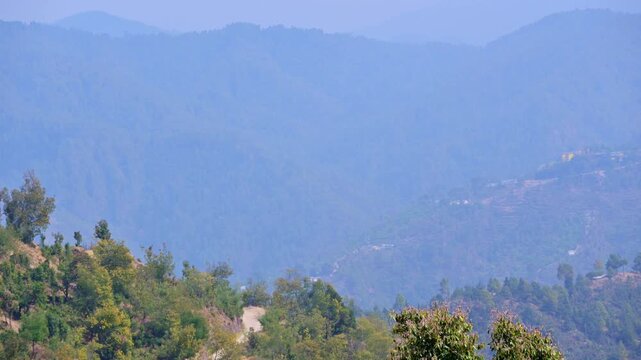 Time-lapse view of Mukteshwar, Uttarakhand, India, highlighting soft sun rays cascading through mist over layered forested hills, winding dirt path and scattered hillside homes emerging through haze.