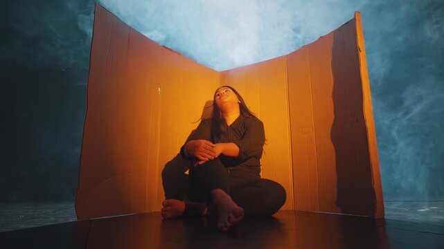 A woman sits alone in a conceptual corner made of cardboard, illuminated by warm light against a contrasting cool blue smoky background. Symbolizes isolation, poverty, or mental barriers.