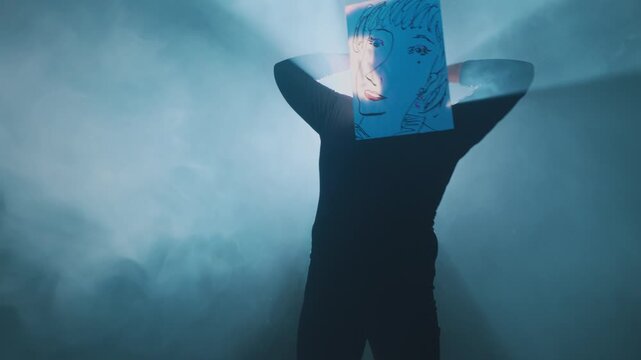 A dancer wearing a hand-drawn female face mask performs fluid, expressive arm movements in a cinematic studio setting with thick blue smoke and dramatic backlighting.