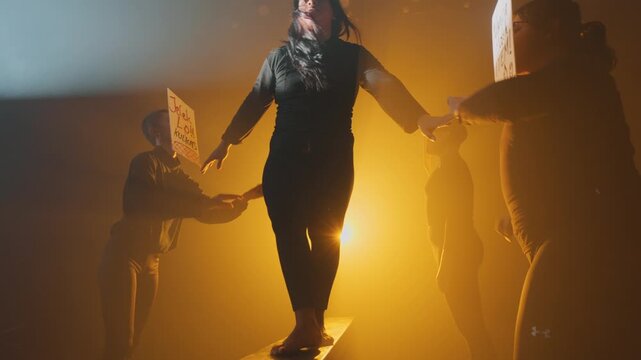 Dramatic low angle of a woman walking a narrow plank on a stage filled with orange smoke. She is hounded by anonymous figures holding abusive text signs, symbolizing toxic judgment and anxiety.
