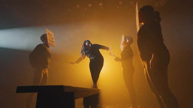 A woman struggles to balance on a wooden beam while masked figures with insulting signs reach out to grab her. A theatrical representation of navigating life under societal pressure and bullying.