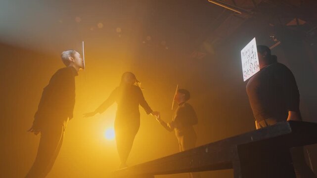 A female actor maintains her balance on a raised platform amidst a dramatic, smoky orange stage. Faceless actors encircle her holding derogatory signs, illustrating a mental health struggle.