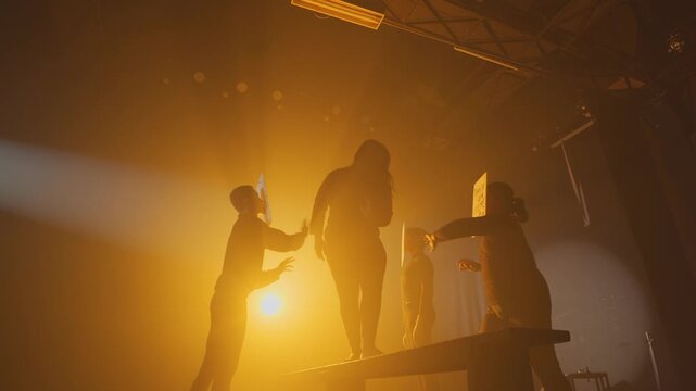 High-contrast, theatrical lighting illuminates a woman balancing on a beam in a foggy studio. She is confronted by anonymous actors holding signs with insults, representing societal pressure.