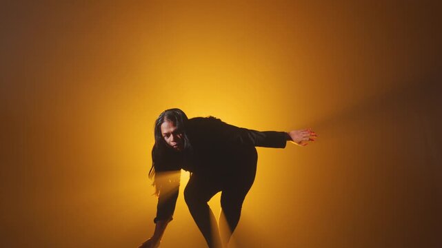 A woman in dark clothing performs a slow, expressive contemporary dance movement, rising from a crouched position on a stage bathed in warm, glowing orange light and atmospheric smoke.