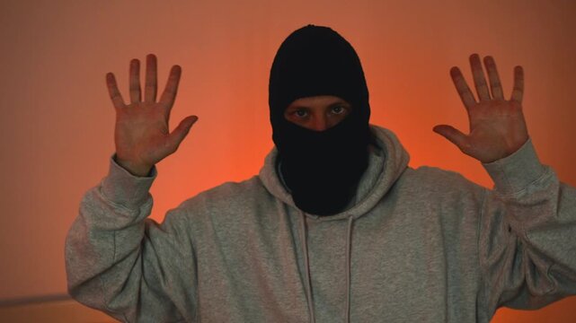 A man in a balaclava surrenders to the police, a law enforcement concept.
Police lights illuminate the room where the criminal is standing, and he surrenders to the cops, realizing he has no choice.