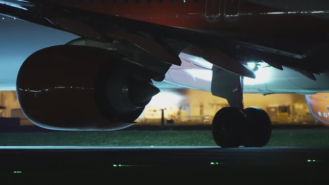 Low angle ground level view of commercial airliner jet engine nacelle and main landing gear wheel rolling on taxiway at night, warm yellow terminal light glowing in background, green taxiway light on