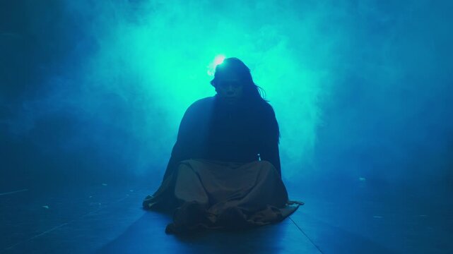 A mysterious person sits crossed-legged on the floor in silhouette, illuminated by a strong blue backlight and dense smoke. Evokes meditation and mystery.