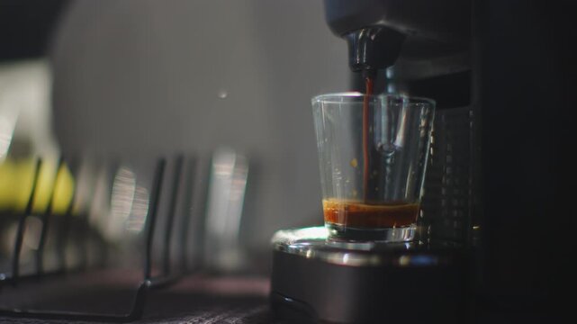 Dark espresso machine brewing a rich espresso shot flowing into a clear glass cup in slow motion, golden brown crema visible, blurred row of glass and yellow light in background