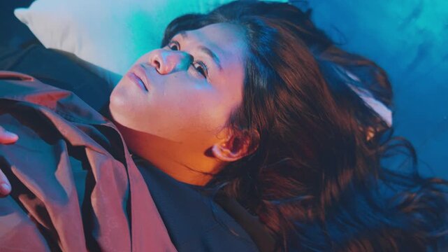 Close-up of a young woman in bed with dramatic blue and orange light. She squeezes her eyes shut before opening them with an intense, contemplative stare.