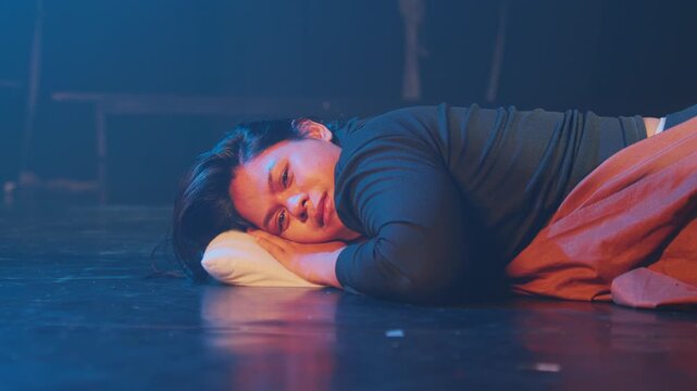 A medium shot of a young woman resting her head on a small pillow on a dark stage floor. She looks softly at the camera with a subtle, bittersweet smile in a blue foggy atmosphere.