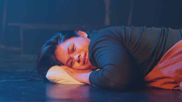 Cinematic shot of a woman lying down on a stage floor with a pillow. She looks toward the camera with a soft, affectionate, or nostalgic expression amidst a moody blue and red lighting setup.