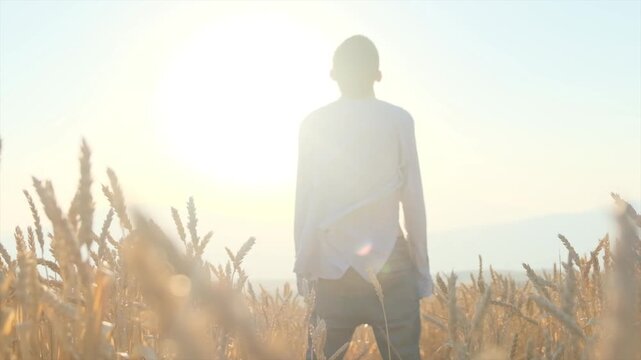Young man stands alone in unbuttoned shirt in wheat field at sunset and tilts head to sky looking at sun. Wind moves shirt of man standing alone among ripe wheat ears. Meditation calm, hope and prayer