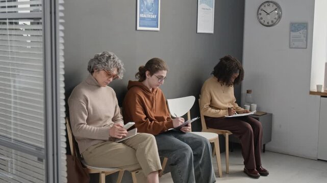 Group of young and mature Caucasian and Black patients filling in forms and using smartphone while sitting on chairs in waiting room