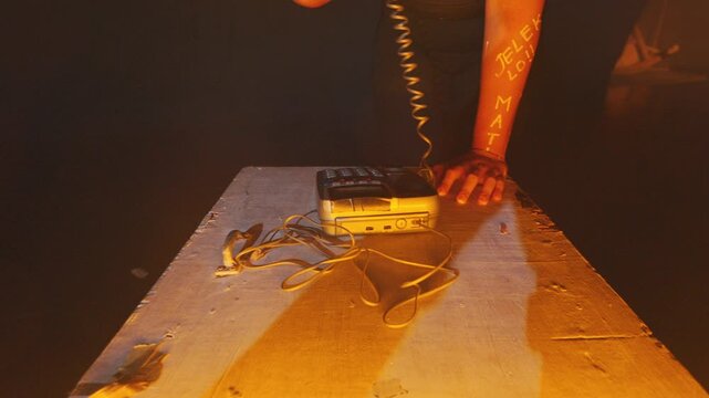 A suspenseful top-down shot of a hand with writing on the skin hovering over an old landline phone. The scene is lit by a striking, moody orange light in a dark room.