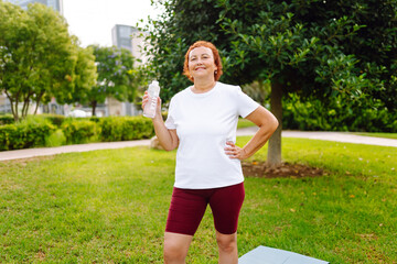 Elderly woman in athletic clothing drinks water from bottle in a sunny park. Athletic adult stands on yoga mat, quenching thirst after workout. Concept of health, sports, and beverages. © maxbelchenko