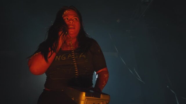 A woman covered in written insults holds an entire vintage telephone, looking furiously into the distance. She is lit by a harsh, dramatic spotlight in a smoky room.