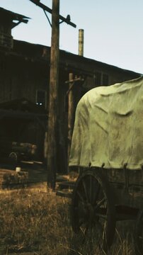 A wagon stands in a dry field close to a wooden structure. The wagon is covered with a piece of cloth. The sun shines on the scene, creating shadows on the ground.