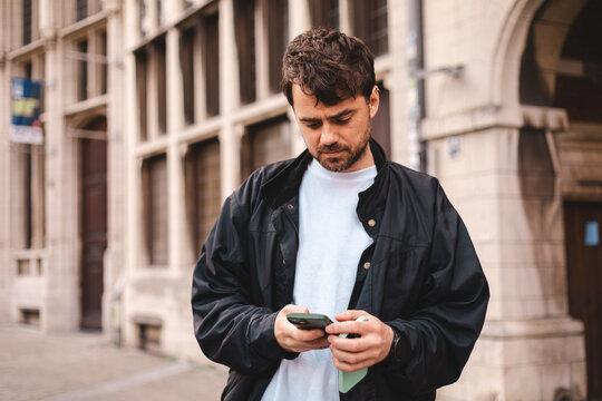 Young Focused Caucasian Man in a Black Jacket Outdoors, Concentration on Smartphone, Holding Two Phones and Fumbling to Attach a Case in a City Street