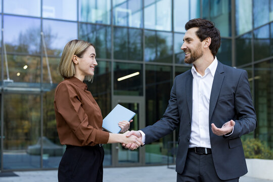 Two smiling business professionals are shaking hands, confirming a successful agreement and partnership outside a modern office building, symbolizing deal closing and collaboration