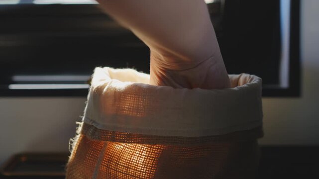 Hand slowly pulling a loaf of bread from a rustic burlap sack in slow motion, flour dust particle floating visible in warm backlight, dark blurred kitchen background