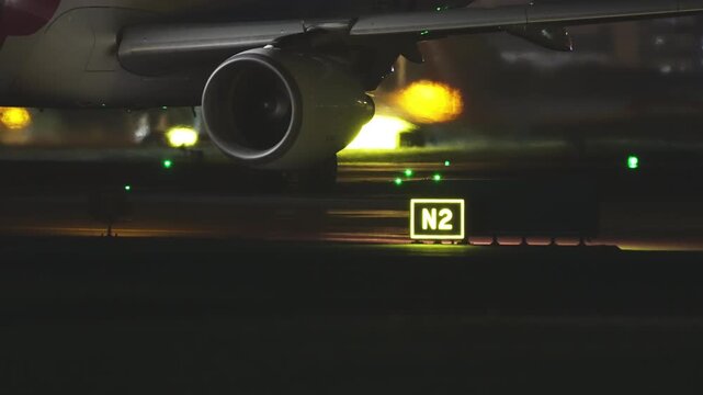 Low angle view of commercial airliner engine underside and wing passing illuminated yellow taxiway sign N2 at night, green taxiway centerline light stretching across dark tarmac, warm light glow on