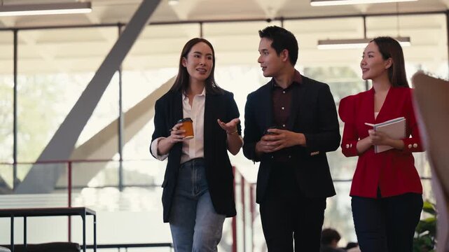 Group of three young Asian businesspeople walking through a modern office, happily chatting and holding coffee cups during a break