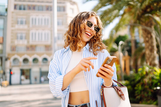 Smiling woman stands on a city street with a phone among palm trees. The woman is texting on phone and enjoying stroll through square on sunny day. Concepts: urban, beauty, blog.