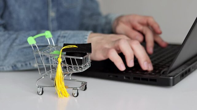 Mini shopping cart with graduation cap beside a laptop, symbolizing online education investment, e-learning costs, tuition fees, and digital learning purchases.
