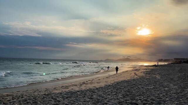 Dramatic sunset sky over seaside with waves as a woman emerges from the silhouettes, jogging toward the foreground in Brazil Rio de Janeiro Barra Tijuca neighborhood extreme long shot