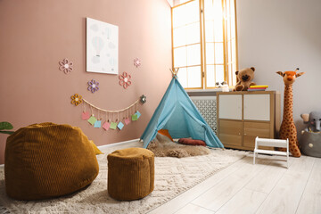 Colorful paper notes and beautiful decor hanging on beige wall near toy wigwam in stylish children's room with chest of drawers and poufs. Interior design © New Africa