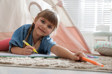 Little girl drawing in sketchbook near toy wigwam at home © New Africa