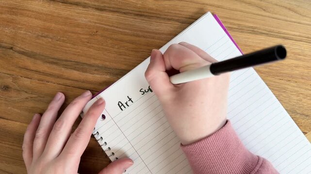 Close up of hands writing &ldquo;art supplies needed&rdquo; on lined notebook paper, creative planning and shopping list concept for artists, natural light overhead shot on wooden table, 4K video