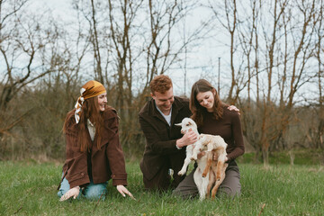 Fototapeta premium Cheerful group outdoors with a young goat kid, friends sharing a gentle moment on lush grass, candid interaction, natural light, relaxed mood, countryside setting