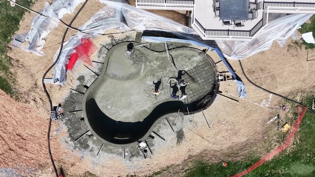 Top-down aerial drone view of workers applying gunite to backyard swimming pool under construction in suburban home in USA, showing landscaping, construction process and residential setting in sun.