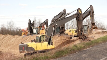 Rząd nowoczesnych koparek gąsienicowych stoi na placu budowy. Potężne, żółte maszyny z długimi ramionami są gotowe do robót ziemnych. W tle widać drzewa i błękitne niebo z delikatnymi chmurami © tomo81