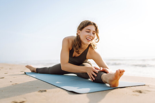 A woman in athletic clothing stretches on a yoga mat on the beach. A young woman meditates and enjoys a workout by the sea. Concepts of freedom, enjoyment, and meditation.