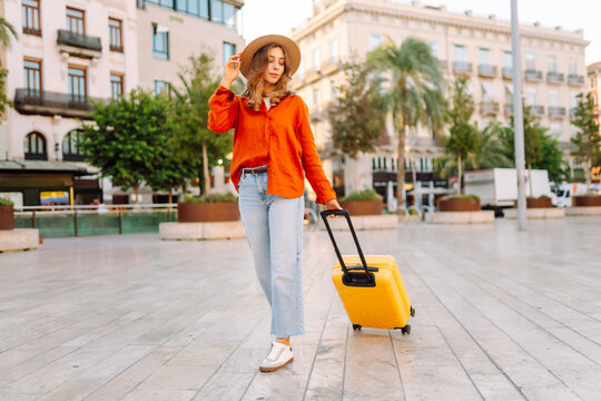 Young traveler wearing hat and carrying small suitcase stands in sunny square. Woman with backpack admires architecture while strolling through city. Concepts for travel, tourism, weekends.