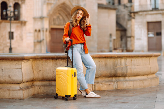 Young traveler wearing hat and carrying small suitcase stands in sunny square. Woman with backpack admires architecture while strolling through city. Concepts for travel, tourism, weekends.