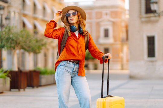 Young traveler wearing hat and carrying small suitcase stands in sunny square. Woman with backpack admires architecture while strolling through city. Concepts for travel, tourism, weekends.