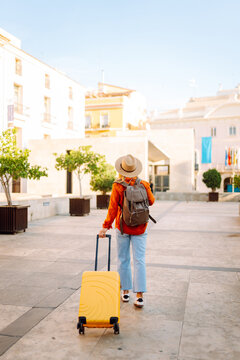 Young traveler wearing hat and carrying small suitcase stands in sunny square. Woman with backpack admires architecture while strolling through city. Concepts for travel, tourism, weekends.