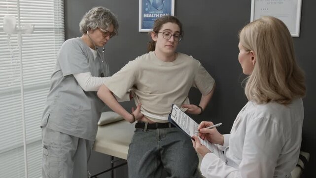 Medium shot of Caucasian teenage boy with long hair undergoing medical examination in modern hospital, doctors checking his heart and lungs with stethoscope and writing notes