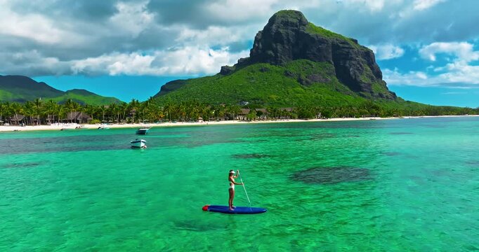 Aerial view of woman in white swimsuite standing on sup board with paddle on crystal turquoise water with coral reef on the tropical island in Indian ocean. Active water sport. Mauritius island