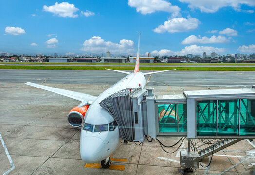 Commercial airplane docked at a boarding bridge at a Brazilian airport. View of the aircraft nose, engine, and jetway against a blue sky and city skyline.